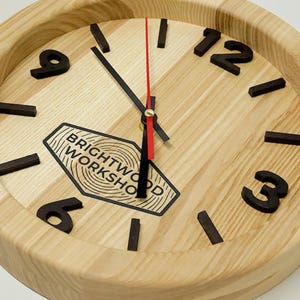 Puede incluir: Un reloj de pared redondo hecho de madera clara con veta visible. Presenta números negros en relieve para el 3, 6, 9 y 12, manecillas negras para las horas y minutos, y un segundero rojo fino. Un logotipo negro dice BRIGHTWOOD WORKSHOP.