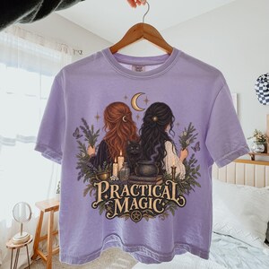 May include: A light purple t-shirt featuring a graphic illustration of two figures with long hair, a black cat, candles, a cauldron, and foliage. The text "PRACTICAL MAGIC" is printed in a decorative font below the figures, with a small pentagram symbol.