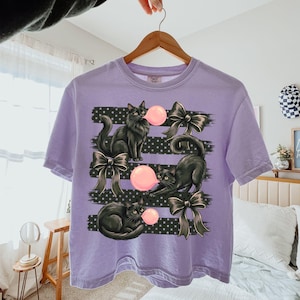May include: A light purple t-shirt featuring a graphic print of three black cats, each blowing a pink bubblegum bubble. The design includes black horizontal stripes with white polka dots and three black bows interspersed among the cats.