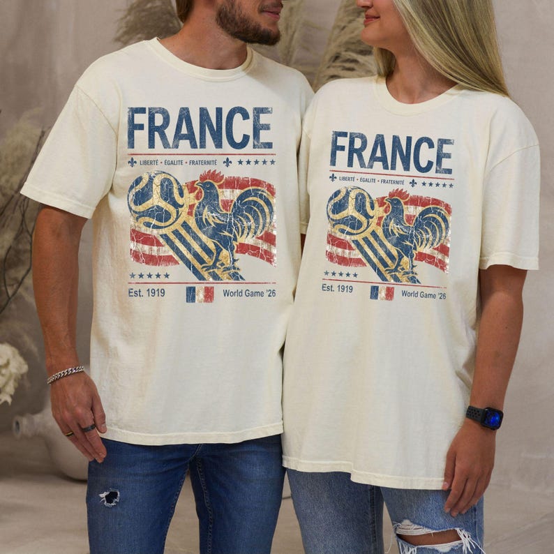 May include: Off-white short-sleeve t-shirts featuring a distressed graphic of a rooster, a soccer ball, and the French flag. The text reads FRANCE, LIBERT&Eacute; &Eacute;GALIT&Eacute; FRATERNIT&Eacute;, Est. 1919, and World Game '26, with five stars above and below the graphic.