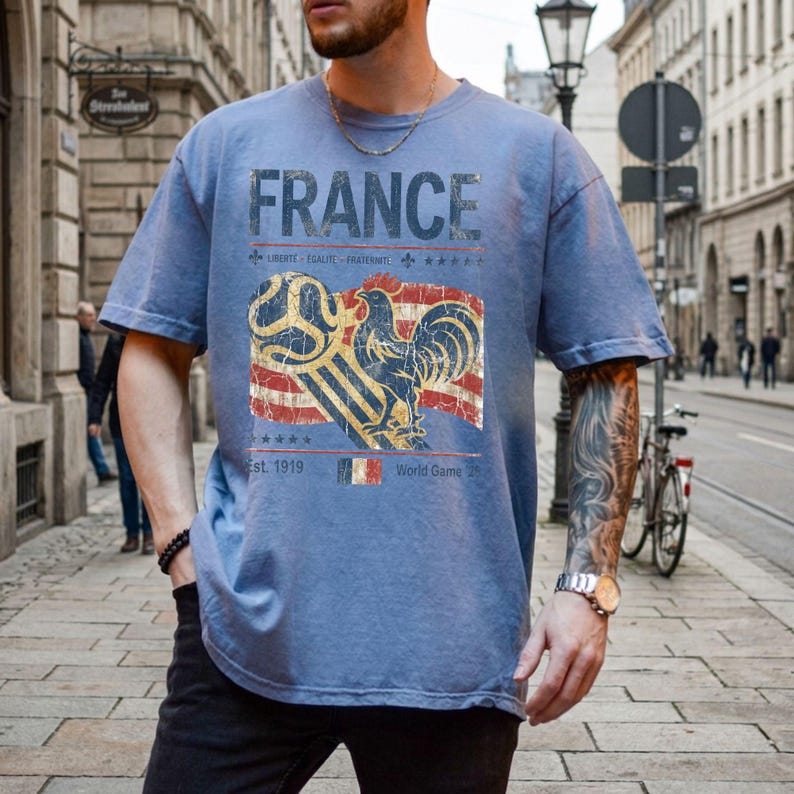 May include: A dusty blue short-sleeve t-shirt featuring a distressed graphic print that reads FRANCE, LIBERTE EGALITE FRATERNITE, Est. 1919, and World Game. The design includes a stylized rooster, a soccer ball, and a French flag motif.