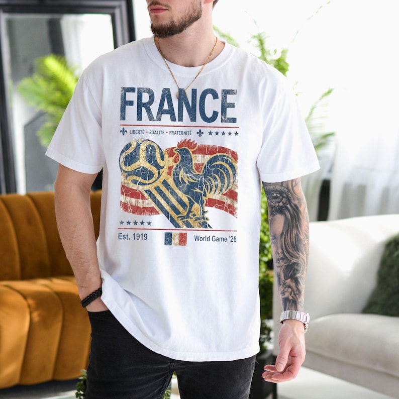 May include: A white short-sleeve t-shirt featuring a graphic with the text FRANCE, LIBERT&Eacute; &Eacute;GALIT&Eacute; FRATERNIT&Eacute;, Est. 1919, and World Game '26. The design includes a blue rooster, a soccer ball, a red, white, and blue flag, and five stars.