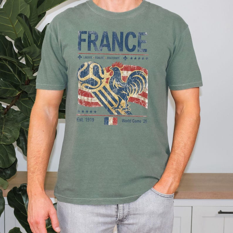 May include: A sage green t-shirt featuring a distressed graphic with the text FRANCE, LIBERT&Eacute;-&Eacute;GALIT&Eacute;-FRATERNIT&Eacute;, Est. 1919, and World Game '26. The design includes a blue rooster, a soccer ball, a striped flag, and a small French flag icon.