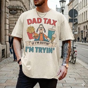 May include: Beige t-shirt with the text "DAD TAX" above a cartoon character holding a burger, fries, a drink, and an ice cream cone. The text "IF I'M BUYIN' I'M TRYIN'" is below the cartoon.