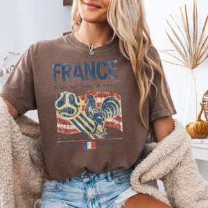 May include: A brown short-sleeve t-shirt featuring the text FRANCE in blue letters. Below the text is a graphic of a rooster and a soccer ball over a red, white, and blue flag design. The print has a distressed, vintage-style texture.