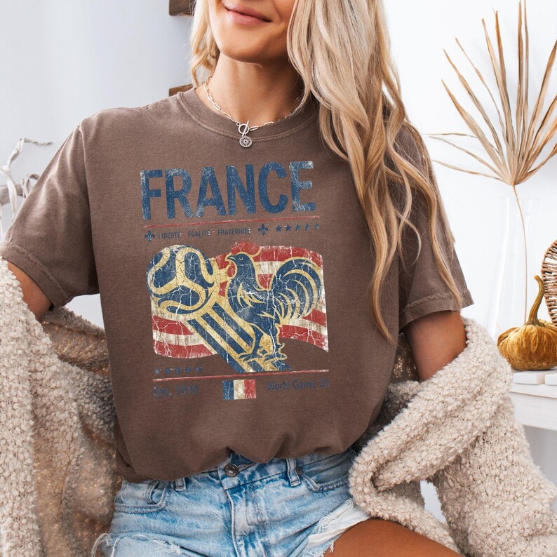 May include: A brown short-sleeve t-shirt featuring the text FRANCE in blue letters. Below the text is a graphic of a rooster and a soccer ball over a red, white, and blue flag design. The print has a distressed, vintage-style texture.