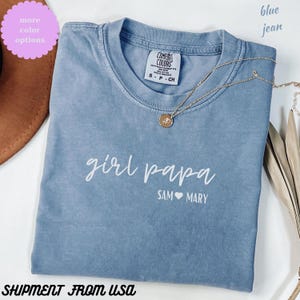 May include: A blue jean Comfort Colors t-shirt with the words "girl papa" and "SAM MARY" printed in white. A gold necklace with a pendant rests on the shirt. The image also includes a pink circle with the words "more color options".