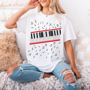 May include: White t-shirt featuring a piano keyboard design with black and white keys and a red border. Black musical notes are scattered around the keyboard. The shirt is made of a soft material.