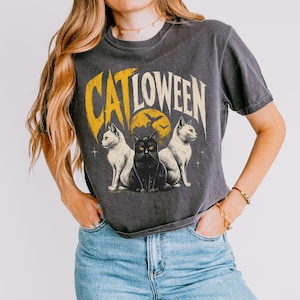 May include: A charcoal gray short-sleeve t-shirt featuring a graphic print with the text CATLOWEEN in yellow. The design includes two white cats and one central black cat sitting in front of a yellow moon with silhouettes of flying bats.