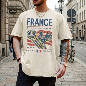 May include: A beige short-sleeve t-shirt featuring a distressed graphic print. The design includes the word FRANCE, a rooster, a soccer ball, a French flag, and the text LIBERTE EGALITE FRATERNITE, Est. 1919, and World Game 26 in blue and red.
