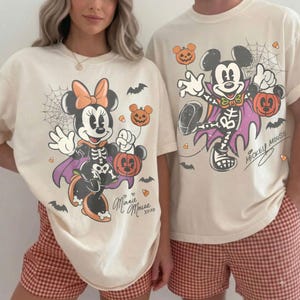May include: Two cream-colored t-shirts featuring Halloween-themed graphics. One shirt displays Minnie Mouse in a skeleton costume with a pumpkin and spiderweb, labeled Minnie Mouse XOXO. The other shows Mickey Mouse in a skeleton cape with a pumpkin.