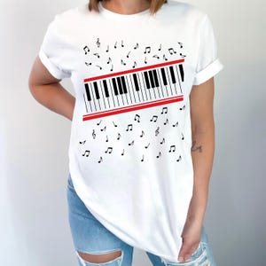 May include: White t-shirt featuring a piano keyboard design with a red stripe and musical notes. The black and white keyboard is the focal point, with musical notes scattered around it. A casual, music-themed graphic tee.