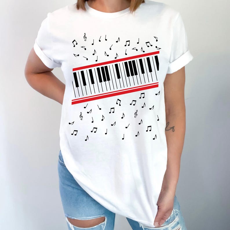 May include: White t-shirt featuring a piano keyboard design with a red stripe and musical notes. The black and white keyboard is the focal point, with musical notes scattered around it. A casual, music-themed graphic tee.