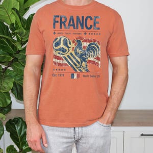 May include: A terracotta-colored short-sleeve t-shirt featuring a distressed graphic print. The design includes the text FRANCE, LIBERTE EGALITE FRATERNITE, Est. 1919, and World Game 26, alongside a rooster, a soccer ball, and a flag motif.