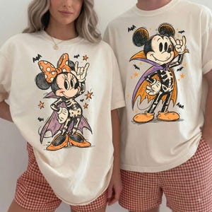 May include: Two off-white t-shirts featuring graphic prints of cartoon characters dressed in skeleton costumes and capes. One shirt displays a character with a polka-dot bow, while the other shows a character with ears, both surrounded by small bats and stars.