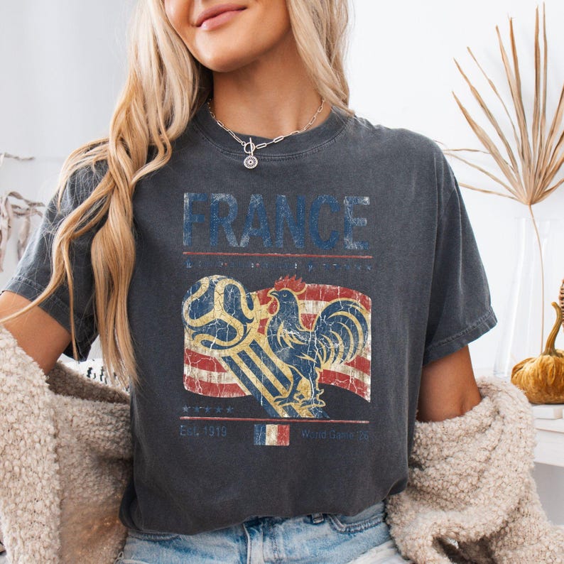 May include: A charcoal gray t-shirt featuring a distressed graphic print with the word FRANCE in blue, a soccer ball, a rooster, and a striped flag background. The design includes the text Est. 1919 and a small French flag icon at the bottom center.