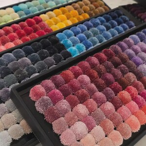 May include: A collection of colorful pom-poms arranged in rows within black trays. The pom-poms display a wide spectrum of colors, including shades of blue, green, yellow, red, and purple. The arrangement creates a visually appealing display of textures and hues.