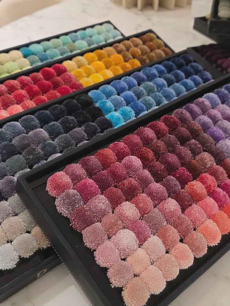 May include: A collection of colorful pom-poms arranged in rows within black trays. The pom-poms display a wide spectrum of colors, including shades of blue, green, yellow, red, and purple. The arrangement creates a visually appealing display of textures and hues.