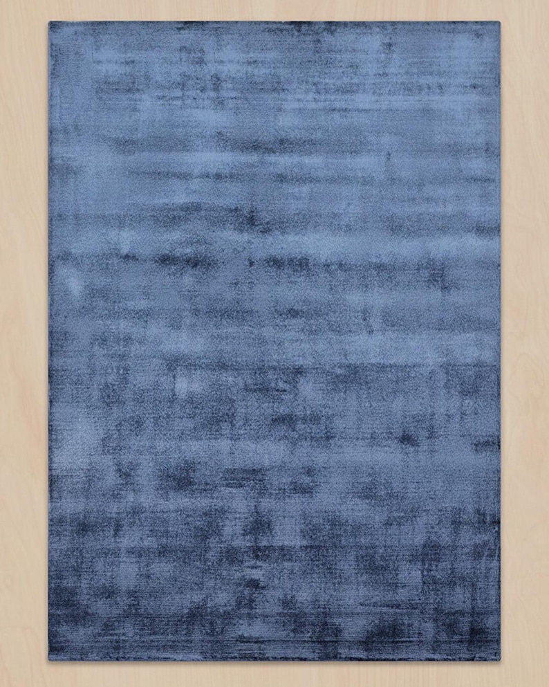 May include: A rectangular, blue area rug with a textured appearance. The rug has a gradient effect, transitioning from a lighter blue at the top to a darker blue at the bottom. The rug is placed on a light-colored wooden floor.
