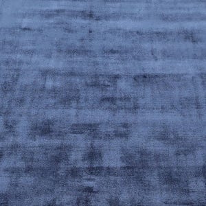 May include: A close-up view of a blue rug with a soft, velvety texture. The rug's color is a deep, muted blue, with subtle variations in tone that create a sense of depth and dimension. The rug appears to be made of a plush material.