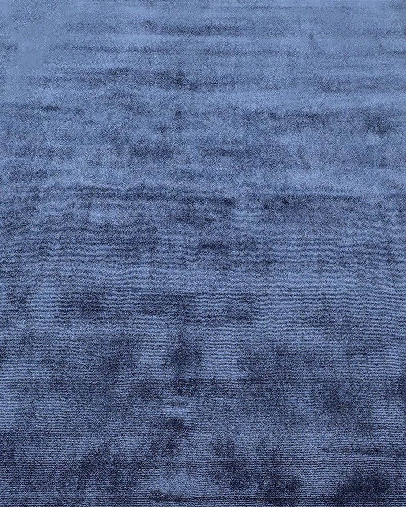 May include: A close-up view of a blue rug with a soft, velvety texture. The rug's color is a deep, muted blue, with subtle variations in tone that create a sense of depth and dimension. The rug appears to be made of a plush material.