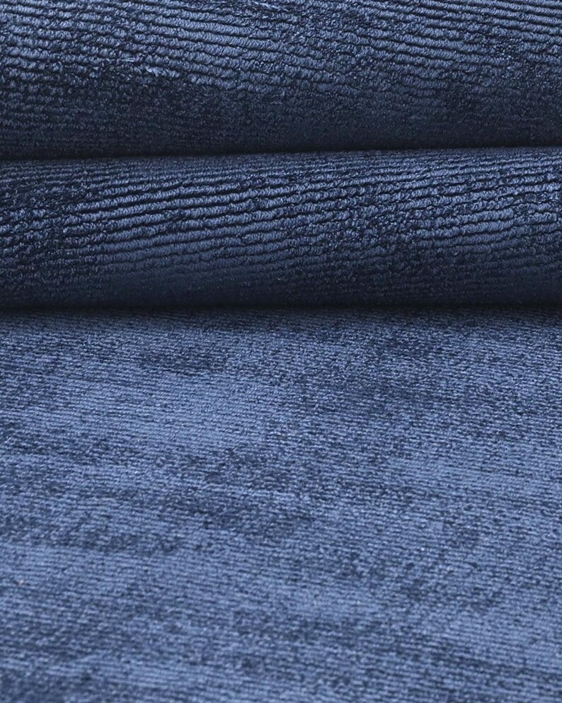 May include: Close-up of a textured, navy blue fabric. The fabric is folded, revealing layers of the material. The texture appears to have a subtle, ribbed pattern, creating a sense of depth and visual interest. The color is a deep, saturated blue.