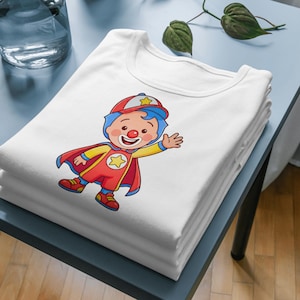 Puede incluir: Una pila de camisetas blancas con una ilustraci&oacute;n de un payaso. El payaso lleva un traje rojo y amarillo con una capa y un sombrero rojos. El personaje tiene pelo azul y una nariz roja.