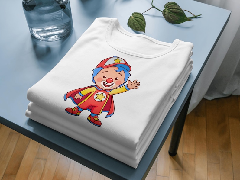Puede incluir: Una pila de camisetas blancas con una ilustraci&oacute;n de un payaso. El payaso lleva un traje rojo y amarillo con una capa y un sombrero rojos. El personaje tiene pelo azul y una nariz roja.