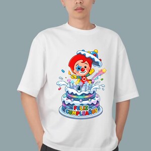 Może przedstawiać: Biały t-shirt z kr&oacute;tkim rękawem z kolorową grafiką przedstawiającą klauna z czerwonymi włosami i niebieską gwiazdką na policzku, wyłaniającego się z wielopoziomowego tortu urodzinowego z zapalonymi świeczkami i napisem "FELIZ CUMPLEA&Ntilde;OS".