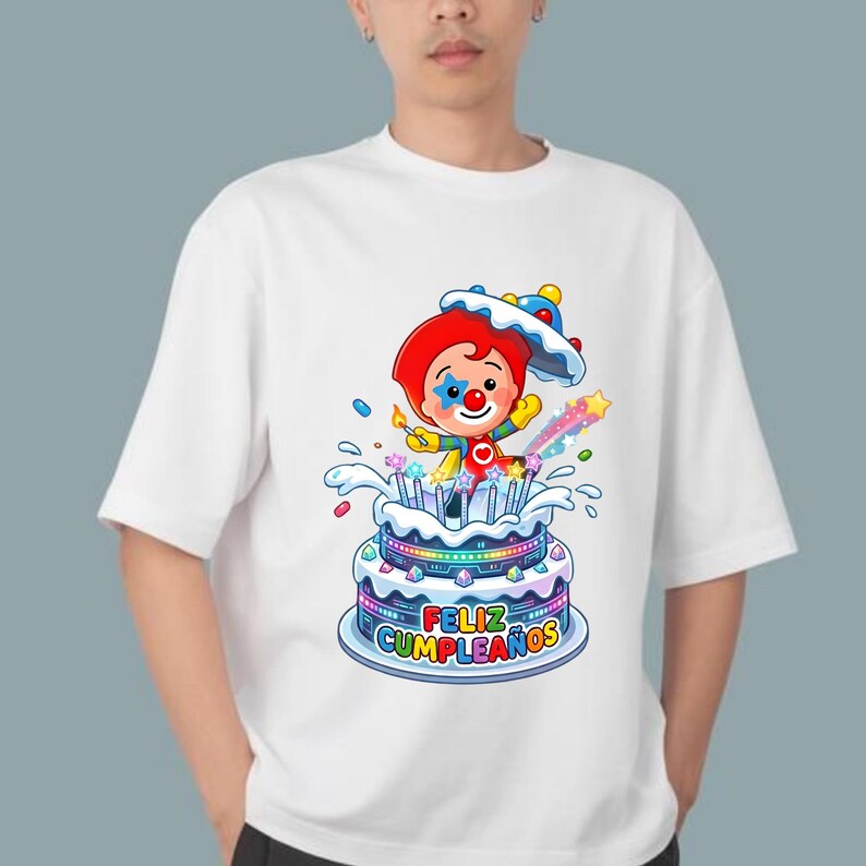 Może przedstawiać: Biały t-shirt z kr&oacute;tkim rękawem z kolorową grafiką przedstawiającą klauna z czerwonymi włosami i niebieską gwiazdką na policzku, wyłaniającego się z wielopoziomowego tortu urodzinowego z zapalonymi świeczkami i napisem "FELIZ CUMPLEA&Ntilde;OS".