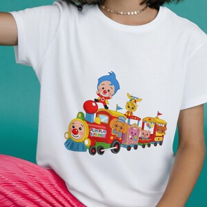 Puede incluir: Camiseta blanca con un gr&aacute;fico colorido de un tren de dibujos animados con personajes. El tren es rojo, amarillo y azul, con el texto "Plim Plim & Friends". La camiseta tiene mangas cortas.