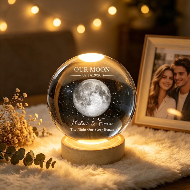 Op de afbeelding: Een heldere glazen bolvormige lamp op een ronde houten voet. Binnenin bevindt zich een gedetailleerde 3D-lasergravure van de maan en sterren. De tekst luidt OUR MOON 02.14.2026, Miles & Fiona, The Night Our Story Began, verlicht door warm licht.