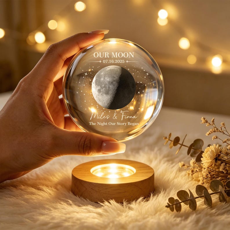Op de afbeelding: Een heldere glazen bol met een gegraveerd ontwerp van een maan en sterren, inclusief de tekst OUR MOON, 07.16.2025, Miles & Fiona en The Night Our Story Began. De bol rust op een ronde, lichtgekleurde houten voet met ge&iuml;ntegreerde warme ledverlichting.