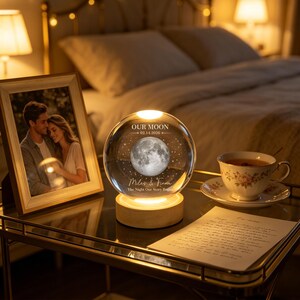 Op de afbeelding: Een heldere glazen bol als nachtlampje op een ronde houten voet, voorzien van een gegraveerde maan, sterren en de tekst "OUR MOON 02.14.2026" en "Miles & Fiona The Night Our Story Began". De bol wordt van onderaf verlicht voor een warme gloed.