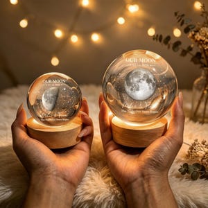 Op de afbeelding: Twee heldere glazen bollen op licht houten voetstukken, elk met een intern lasergegraveerd maan- en sterrenontwerp. De bollen zijn gegraveerd met de tekst OUR MOON, datums, de namen Miles & Fiona en de zin The Night Our Story Began.