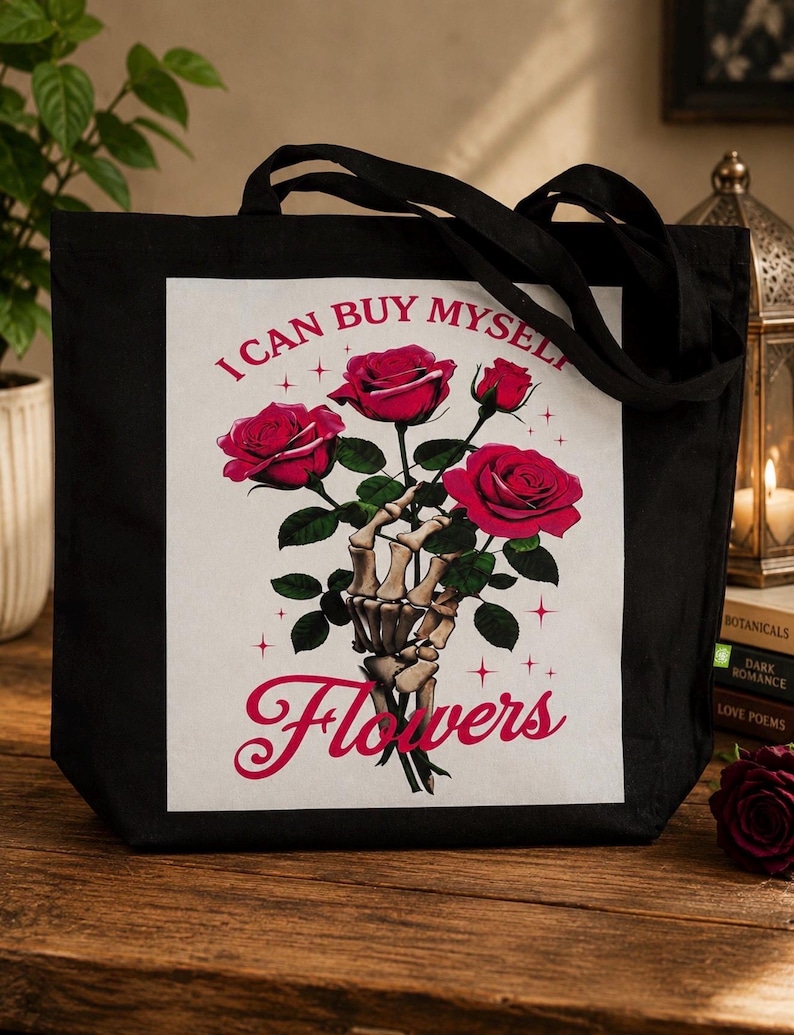 May include: A black tote bag featuring a graphic print of a skeletal hand holding a bouquet of three red roses. The text "I CAN BUY MYSELF" is printed in red above the roses, and the word "Flowers" is printed in a large red script font below.