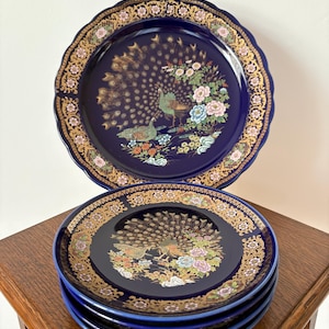 May include: A set of five round ceramic plates with a deep cobalt blue glaze. Each plate features a central illustration of a peacock with a fanned tail, surrounded by colourful floral motifs and a wide, ornate gold filigree border on the rim.