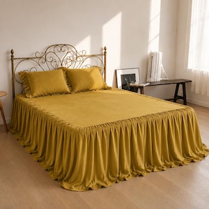 May include: A mustard-yellow bed with a decorative metal headboard. The bed has a matching bedspread and two pillows. The bedspread has a ruffled edge. A small wooden side table with a plant and vase is to the left.