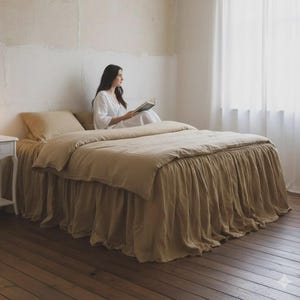 May include: A bedroom scene featuring a bed with a beige linen duvet cover and a ruffled bed skirt. A white nightstand with a lamp sits beside the bed. A person in a white dress is sitting on the bed reading a book.