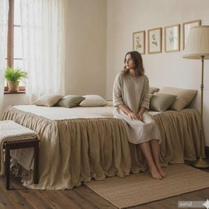 May include: A bedroom scene with a bed featuring a beige bed skirt and pillows. A woman in a beige dress sits on the bed. A woven rug and a bench with a blanket are in front of the bed. The room has a neutral color palette.