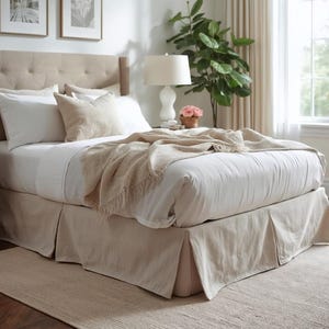 Pode incluir: Uma cama estofada bege com roupa de cama branca, almofadas e uma manta com franjas. A cama tem uma saia de cama a condizer. Duas fotos emolduradas estão penduradas na parede acima da cama. Uma lâmpada e uma planta estão à direita da cama.