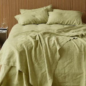 May include: A bed dressed with light green linen bedding, including a fitted sheet, flat sheet, and two pillowcases. The bed is against a wooden panel headboard. A small side table with books and decorative items is visible.