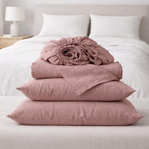 May include: A stack of dusty rose-colored bed linens, including pillowcases, a fitted sheet, and a flat sheet, are arranged on a bench. The bedding is made of a soft, textured fabric. A white bed and pillows are in the background.