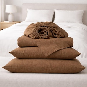 May include: A stack of brown linen bedding, including a fitted sheet, flat sheet, and two pillowcases. The bedding is arranged on a light-colored bench in front of a bed with white linens. The room has a neutral color palette.