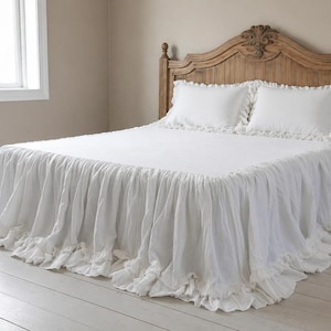 Pode incluir: Uma cama com cabeceira de madeira e roupa de cama branca. A cama tem uma saia de cama branca com babados e duas almofadas brancas com babados. A cabeceira é feita de madeira marrom clara com detalhes esculpidos. O quarto tem uma paleta de cores neutras.