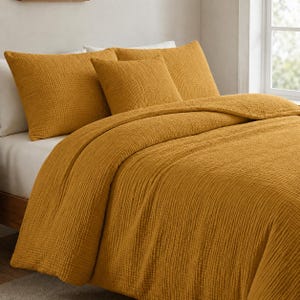Peut inclure: Un ensemble de literie moutarde avec une housse de couette texturée et des taies d'oreiller assorties. L'ensemble est fait d'un tissu doux et tissé. Le lit est fait avec des draps blancs. L'ensemble est présenté sur un cadre de lit en bois.