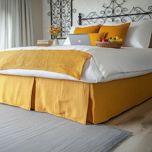 Pode incluir: Uma cama com lençóis brancos, almofadas amarelo mostarda e uma saia de cama a condizer. Um laptop e uma cesta de frutas estão na cama. A cabeceira é de ferro forjado preto e o quarto tem uma paleta de cores neutras.