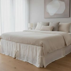 May include: A bed with a neutral-toned duvet cover and matching pillows. The bed has a white bed skirt with tie details. The room features light-colored walls, sheer curtains, and a wooden floor. A painting hangs above the bed.