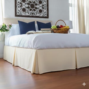 Pode incluir: Uma cama com uma saia de cama plissada creme, lençóis brancos e almofadas azul marinho. Uma cesta de vime cheia de frutas está sobre a cama. Uma decoração de parede e um vaso de flores também são visíveis.