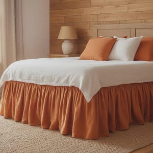 May include: A bed with a white duvet and an orange bed skirt. The bed has orange and white pillows. The room features a wooden headboard, a wooden wall, and a beige rug. A lamp and a window with curtains are also visible.